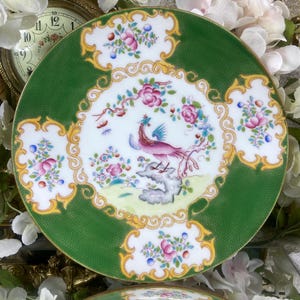 May include: A green and white porcelain plate with a gold trim and a detailed floral design featuring a pink and purple bird. The plate has a scalloped edge and a raised centre.