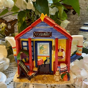 Può includere: Una teiera in ceramica colorata a forma di casa sulla spiaggia con tetto rosso, pareti blu e finiture gialle. La teiera ha un cartello che dice "Seaview" e presenta dettagli a tema spiaggia come un ombrellone da spiaggia, sedie a sdraio e un castello di sabbia.