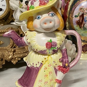 Puede incluir: Una tetera de cerámica caprichosa con forma de cerdo, que lleva un sombrero amarillo adornado con flores. El cerdo está vestido con un vestido amarillo y morado, con guantes a juego y un parasol rojo. La tetera tiene un diseño pintado a mano.