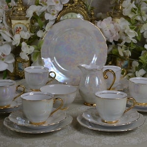 Puede incluir: Un juego de té vintage con acabado nacarado, que incluye tazas, platillos, una crema, un azucarero y un plato grande. El juego tiene adornos y asas doradas, y está dispuesto sobre una mesa con decoraciones florales.
