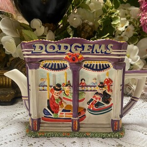 May include: A ceramic teapot shaped like a fairground ride with the words "DODGEMS" on the top. The teapot is decorated with clowns, cars, and other fairground elements.