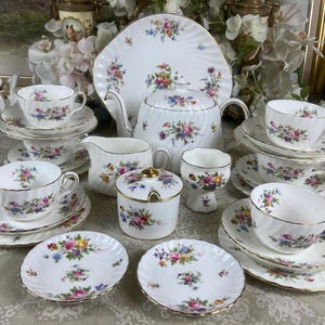 May include: A white porcelain tea set featuring a delicate floral pattern with gold trim. The set includes a teapot, creamer, sugar bowl, cups, saucers, and plates, all decorated with colourful bouquets of flowers on a ribbed, scalloped surface.