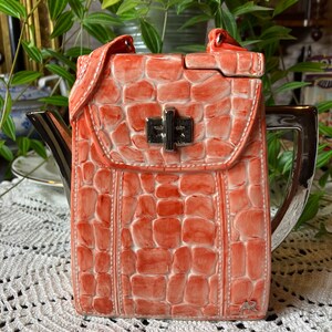 May include: A ceramic teapot shaped like a handbag with a crocodile skin pattern. The teapot is orange with a silver handle and spout. The lid has a silver clasp.