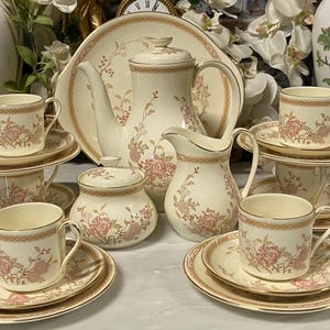 Könnte beinhalten: Cremefarbenes Teeservice mit rosa Blumenmustern. Das Set umfasst Tassen, Untertassen, eine Teekanne, einen Sahnekännchen, eine Zuckerdose und eine Servierplatte. Goldfarbene Verzierungen akzentuieren die Ränder jedes Stücks und schaffen eine elegante Ästhetik.