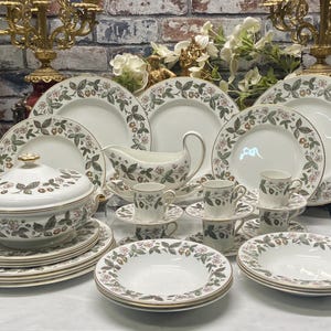 Op de afbeelding: Een crèmekleurige porseleinen serviescollectie met een herhalend patroon van groene bladeren, kleine rode aardbeien en roze bloesems. De set bevat diverse borden, een terrine met deksel, een sauskom en verschillende kleine kopjes.