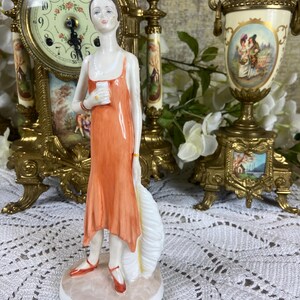 Può includere: Statuina di porcellana di una donna in abito arancione, con un ventaglio bianco. La statuina ha scarpe rosse e una collana di perle. Un orologio dorato e urne decorative sono sullo sfondo.