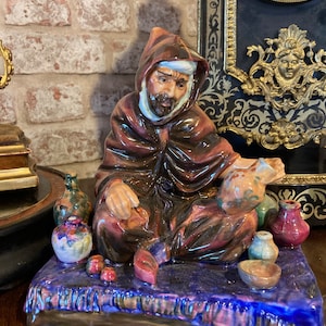 Puede incluir: Una figurita de cerámica de un hombre con una túnica marrón y un turbante blanco, sentado en una alfombra azul y morada. Está sosteniendo una olla de cerámica marrón y blanca. Hay varias ollas de cerámica más pequeñas y fruta en la alfombra.