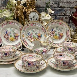 May include: A porcelain tea set featuring three plates, three teacups with matching saucers, and a creamer pitcher. Each piece is decorated with a colorful floral and geometric pattern in pink, yellow, blue, and green, finished with gold trim.