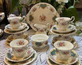 May include: A vintage tea set with floral and gold trim. The set includes teacups, saucers, a serving plate, a creamer, and a sugar bowl. The teacups and saucers are stacked on a white lace tablecloth. The set is displayed in front of a floral arrangement.
