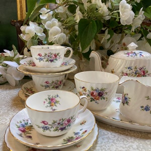 May include: A classic porcelain tea set, including cups, saucers, a milk jug, and a teapot. The white china is decorated with colourful floral designs in pink, purple, blue, and yellow, with gold edging. The set is displayed on a patterned cloth.