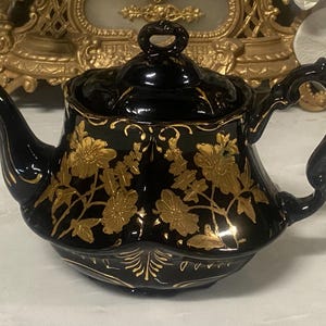May include: A black ceramic teapot with a lid, handle, and spout. The teapot is adorned with gold floral designs. The handle is curved, and the spout is slender. The teapot sits on a white surface.