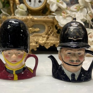 May include: Two novelty teapots. One is a ceramic figure of a British Royal Guard in a red coat and black hat. The other is a police officer in a dark blue uniform and helmet. Both have detailed faces and are decorative.