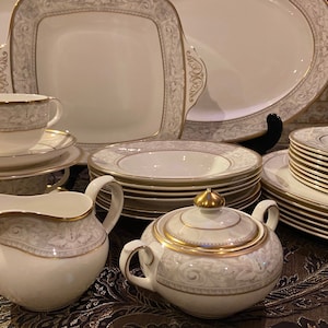 May include: A set of white fine china dinnerware with gold trim and a floral pattern. The set includes plates, bowls, a creamer, a sugar bowl, and a serving platter.