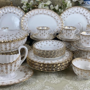 Puede incluir: Un juego de vajilla de porcelana blanca con ribetes dorados y un patrón floral. El juego incluye platos, tazones, tazas, platillos, una tetera, una salsera y una jarra.