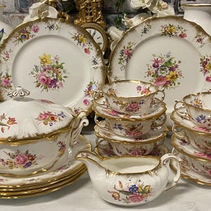 Puede incluir: Juego de vajilla antiguo con diseños florales. El conjunto incluye platos, cuencos, tazas, platillos, una fuente para servir y una jarra para crema. Cada pieza es blanca con ribete dorado y un patrón floral colorido.