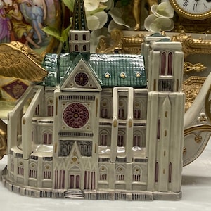 May include: A ceramic teapot shaped like a cathedral with a green roof, cream-coloured walls, and a rose window. The handle is a gold-coloured angel with wings, and the spout is an elongated architectural extension with gold trim and accents.