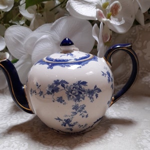 Op de afbeelding: Een witte theepot met een blauw en gouden bloemenmotief. De theepot heeft een blauwe handgreep en een gouden rand.