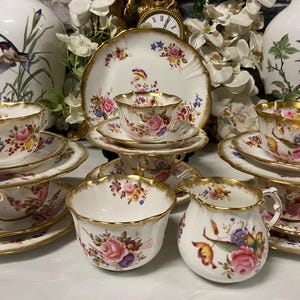 Può includere: Una collezione di tazze da tè, piattini e piatti di servizio in porcellana antica. La porcellana bianca è decorata con motivi floreali rosa, blu e gialli, con bordi dorati. Il set comprende tazze, piattini, ciotole e una lattiera.