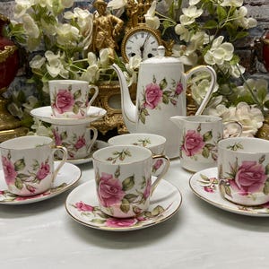 May include: A white porcelain tea set featuring a pink rose floral pattern with gold trim. The set includes a tall teapot, a creamer, and four teacups with matching saucers, all decorated with detailed pink roses and green leaves on a white base.