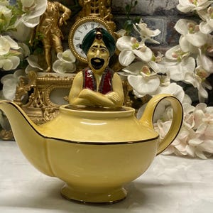 Pode incluir: Uma chaleira amarela com uma figura de gênio no topo. A chaleira tem um bico e uma alça curvos, com uma borda marrom escura. O gênio está usando um turbante verde, um colete vermelho e tem uma barba preta. O fundo inclui flores brancas e um relógio decorativo.
