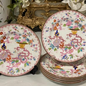 Könnte beinhalten: Satz dekorativer Porzellanteller mit floralem und geometrischem Design. Die Teller haben einen weißen Hintergrund mit rosa, blauen, orangefarbenen und gelben Blumenmustern und einem rosa Rand. Die Teller werden auf schwarzen Ständern ausgestellt.