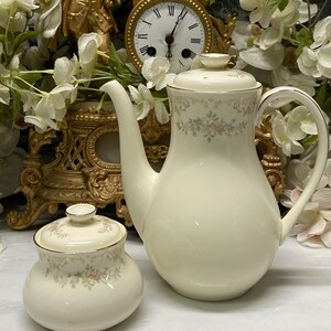 Puede incluir: Una cafetera y un azucarero a juego de porcelana en color crema, ambos con un delicado patrón floral en tonos suaves de rosa, amarillo y verde, con bordes plateados en las tapas, asas y bordes, presentados como un conjunto de dos piezas.