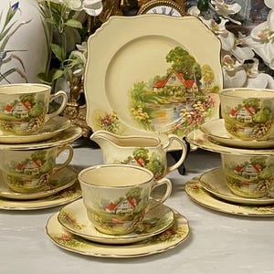 May include: A vintage tea set with a cream background. The set includes teacups, saucers, plates, and a creamer, all decorated with a scenic landscape design featuring a house, trees, and water. Gold trim edges.
