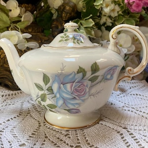 Peut inclure: Une théière blanche avec une bordure dorée et un motif floral composé de roses bleues et violettes. La théière a un bec courbé et une poignée.
