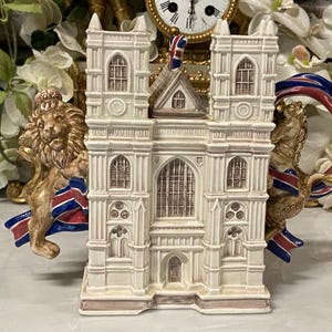 May include: A cream-coloured ceramic teapot in the shape of a cathedral, with detailed architectural features. It has brown accents and a Union Jack flag. The handle is decorated with a golden lion and red, white, and blue ribbons.