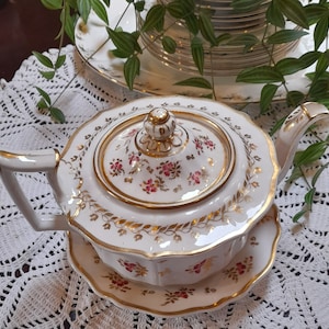 May include: A white porcelain teapot with a gold trim and floral design. The teapot has a lid with a knob and a spout. The teapot is sitting on a matching saucer.