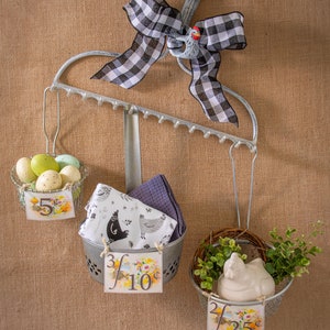 May include: A metal rack with three hanging baskets. The top basket holds a collection of speckled Easter eggs. The middle basket holds a blue and white patterned tea towel. The bottom basket holds a white ceramic chicken figurine with green foliage. Each basket has a price tag attached.