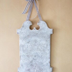 May include: A white wooden sign with a distressed finish hangs from a blue and white striped ribbon. The sign reads "Welcome to Knob Hill Farm".
