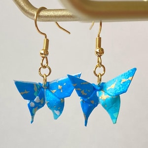 Könnte beinhalten: Ein Paar Origami-Schmetterlingsohrringe. Die blauen Schmetterlinge mit goldenen Akzenten hängen an goldfarbenen Ohrhaken. Ein einzigartiges und künstlerisches Schmuckaccessoire.