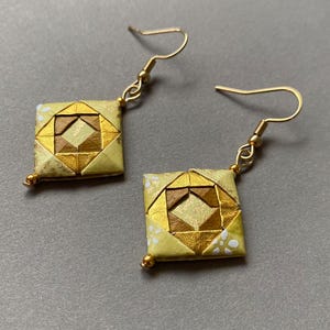 K&ouml;nnte beinhalten: Ein Paar handgefertigte Origami-Ohrringe. Jeder Ohrring hat ein quadratisches Design mit goldenen und braunen geometrischen Mustern. Die Ohrringe haben goldfarbene Ohrhaken und kleine goldene Perlen. Das Papier hat einen hellgelben Hintergrund mit wei&szlig;en Punkten.