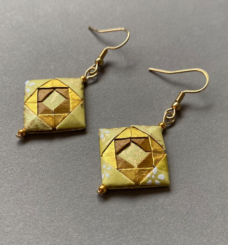 K&ouml;nnte beinhalten: Ein Paar handgefertigte Origami-Ohrringe. Jeder Ohrring hat ein quadratisches Design mit goldenen und braunen geometrischen Mustern. Die Ohrringe haben goldfarbene Ohrhaken und kleine goldene Perlen. Das Papier hat einen hellgelben Hintergrund mit wei&szlig;en Punkten.