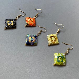 K&ouml;nnte beinhalten: F&uuml;nf Paar quadratische Origami-Ohrringe in verschiedenen Farben, darunter Schwarz, Orange, Marineblau, Gold und Gr&uuml;n. Jeder Ohrring hat einen goldfarbenen Haken und eine kleine goldene Perle am unteren Ende. Die Ohrringe werden auf einer grauen Oberfl&auml;che pr&auml;sentiert.