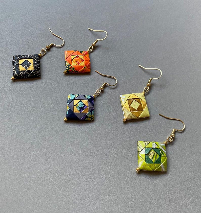 K&ouml;nnte beinhalten: F&uuml;nf Paar quadratische Origami-Ohrringe in verschiedenen Farben, darunter Schwarz, Orange, Marineblau, Gold und Gr&uuml;n. Jeder Ohrring hat einen goldfarbenen Haken und eine kleine goldene Perle am unteren Ende. Die Ohrringe werden auf einer grauen Oberfl&auml;che pr&auml;sentiert.