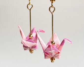 May include: A pair of gold earrings with pink origami cranes hanging from them. The cranes are made of patterned paper and have gold beads at the bottom.