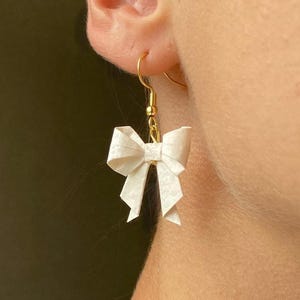 Könnte beinhalten: Nahaufnahme eines einzelnen Ohrrings mit einer weißen Papier-Schleife. Die Schleife hängt an einem goldfarbenen Haken. Der Ohrring ist an einem Ohr befestigt. Die Schleife hat ein strukturiertes Aussehen und besteht aus gefaltetem Papier.