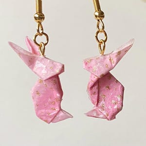 Könnte beinhalten: Ein Paar Origami-Hasenohrringe. Jeder Hase ist aus rosa Papier mit Goldflocken gefertigt und an goldfarbenen Ohrhaken befestigt. Die Ohrringe zeigen ein zartes, handgefertigtes Design und verleihen jedem Outfit einen Hauch von Laune.