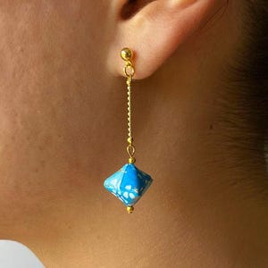 Könnte beinhalten: Ein goldfarbener Ohrring mit einem kleinen Kugelstecker und einer Kette, die eine blau-weiß geometrische Perle hält. Der Ohrring wird am Ohr einer Person gezeigt.