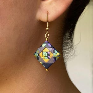 May include: A single gold-colored earring with a geometric, origami-style pendant. The pendant is a diamond shape, featuring a pattern of dark blue, gold, and teal. The earring is attached to a gold-colored hook.