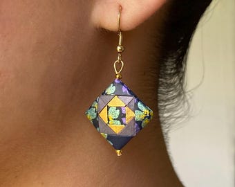 May include: A single gold-colored earring with a geometric, origami-style pendant. The pendant is a diamond shape, featuring a pattern of dark blue, gold, and teal. The earring is attached to a gold-colored hook.