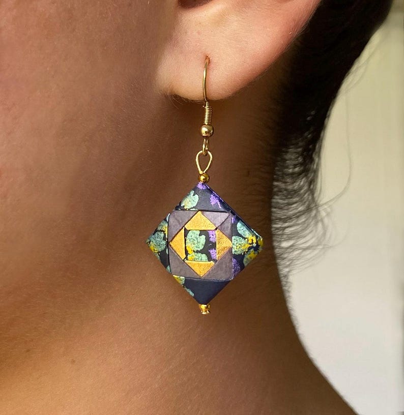 K&ouml;nnte beinhalten: Ein einzelner goldfarbener Ohrring mit einem geometrischen Anh&auml;nger im Origami-Stil. Der Anh&auml;nger hat eine rautenf&ouml;rmige Form mit einem Muster aus Dunkelblau, Gold und T&uuml;rkis. Der Ohrring ist an einem goldfarbenen Haken befestigt.