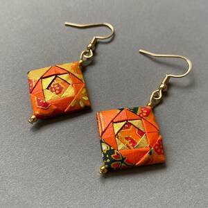 K&ouml;nnte beinhalten: Origami-Ohrringe mit geometrischem Design. Die Ohrringe haben eine quadratische Form mit orangefarbenen, goldenen und roten Mustern. Goldfarbene Ohrhaken und kleine goldene Perlen vervollst&auml;ndigen das Design.