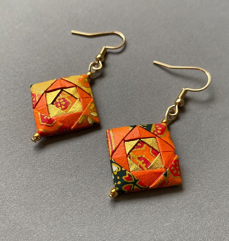 K&ouml;nnte beinhalten: Origami-Ohrringe mit geometrischem Design. Die Ohrringe haben eine quadratische Form mit orangefarbenen, goldenen und roten Mustern. Goldfarbene Ohrhaken und kleine goldene Perlen vervollst&auml;ndigen das Design.