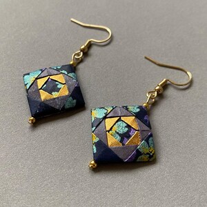 K&ouml;nnte beinhalten: Ein Paar quadratische Origami-Ohrringe mit goldfarbenen Fischhaken-Ohrdr&auml;hten. Die Ohrringe haben ein geometrisches Design mit marineblauen, grauen, goldenen, t&uuml;rkisfarbenen und lila Akzenten. Jeder Ohrring hat eine kleine goldene Perle unten.