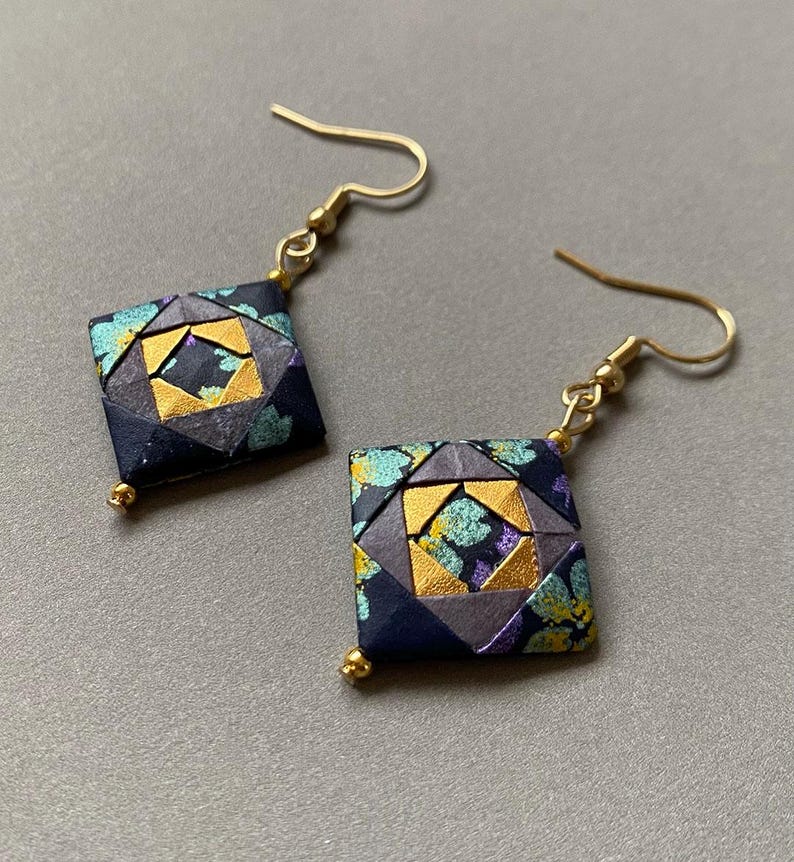 K&ouml;nnte beinhalten: Ein Paar quadratische Origami-Ohrringe mit goldfarbenen Fischhaken-Ohrdr&auml;hten. Die Ohrringe haben ein geometrisches Design mit marineblauen, grauen, goldenen, t&uuml;rkisfarbenen und lila Akzenten. Jeder Ohrring hat eine kleine goldene Perle unten.
