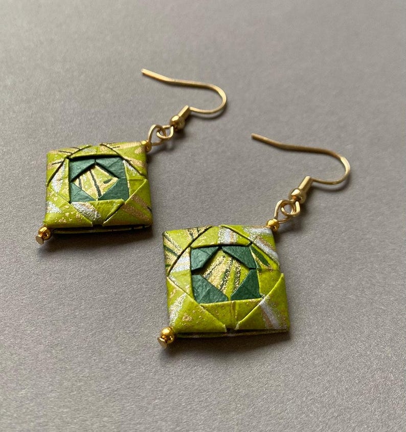 K&ouml;nnte beinhalten: Ein Paar quadratische Origami-Ohrringe mit goldfarbenen Ohrdr&auml;hten und kleinen goldenen Perlen. Die Ohrringe weisen ein geometrisches Design in Gr&uuml;n-, Gold- und Silbert&ouml;nen auf. Die Ohrringe werden auf grauem Hintergrund pr&auml;sentiert.