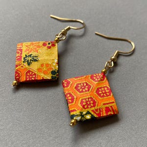 K&ouml;nnte beinhalten: Ein Paar quadratische H&auml;ngeohrringe. Die Ohrringe haben goldfarbene Haken und quadratische Anh&auml;nger mit einem farbenfrohen, gemusterten Design in Orange, Rot und Gold. Jeder Ohrring hat eine kleine goldene Perle unten.